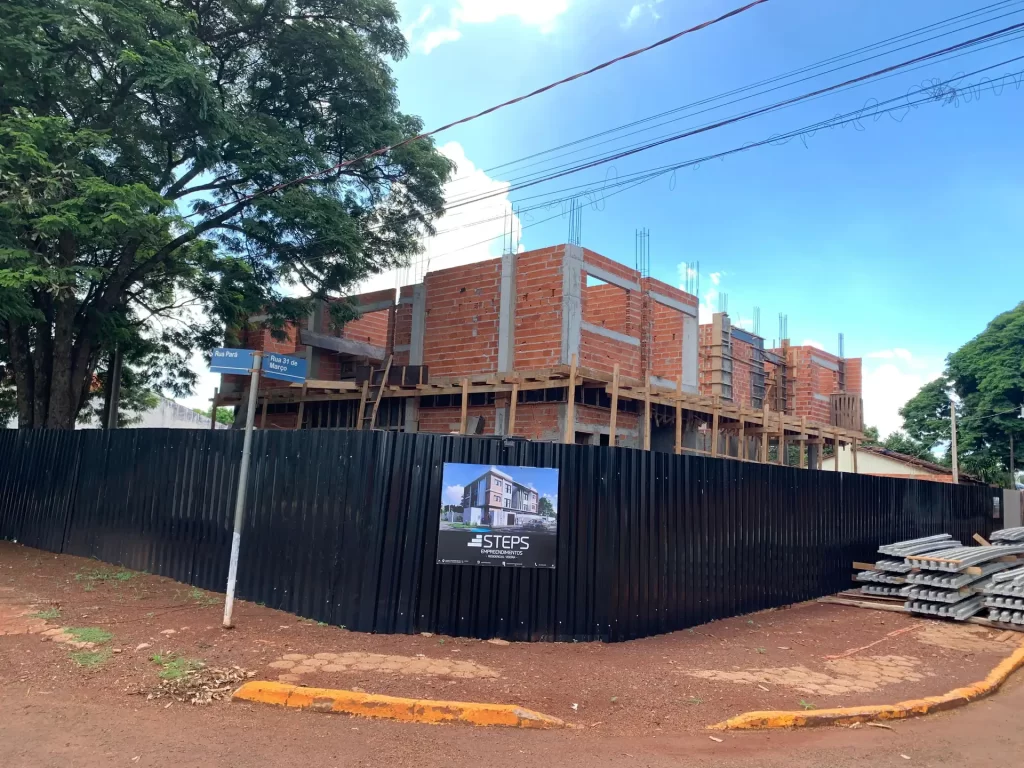 Sobrado em estrutura de tijolo atrás de tapume preto com placa STEPS, obra de esquina