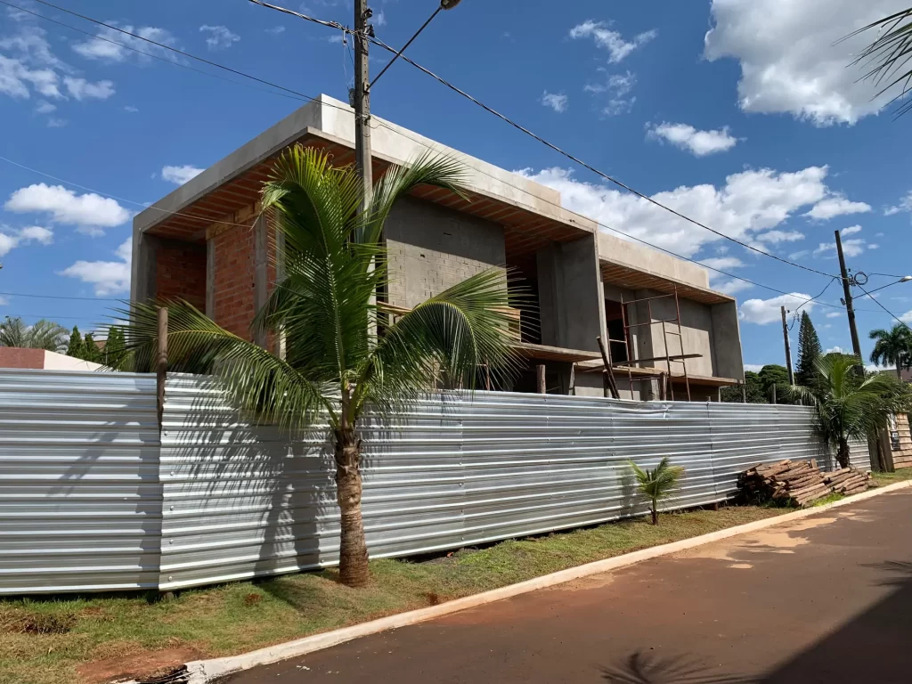 Sobrado de concreto aparente em estrutura atrás de tapume metálico, com coqueiros na frente, obra