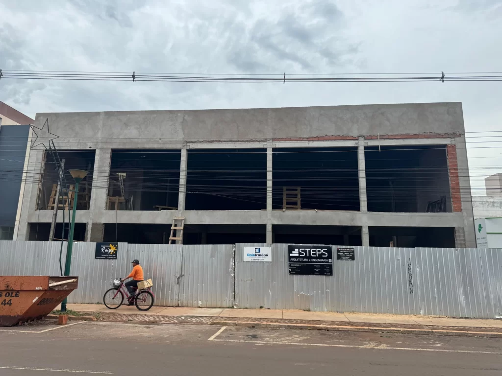 Prédio comercial de concreto em estrutura na avenida atrás de tapume com placas STEPS e Dois Irmãos