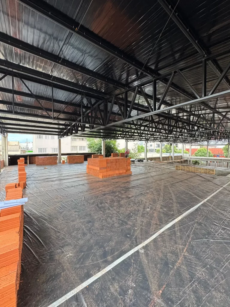 Interior de galpão com cobertura metálica, pilhas de tijolos sobre contrapiso coberto com lona, obra