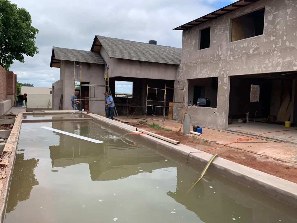 Piscina de concreto recém-concretada diante de sobrado em obra com operários no reboco