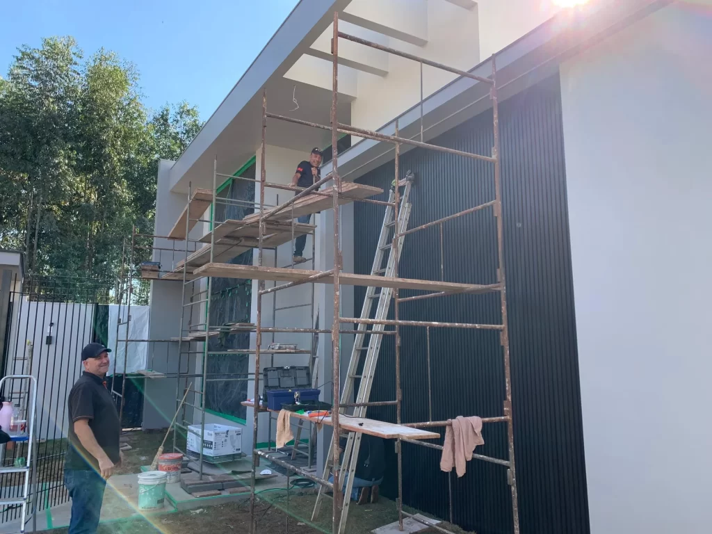 Operários em andaime aplicando revestimento ripado escuro em fachada de residência, obra
