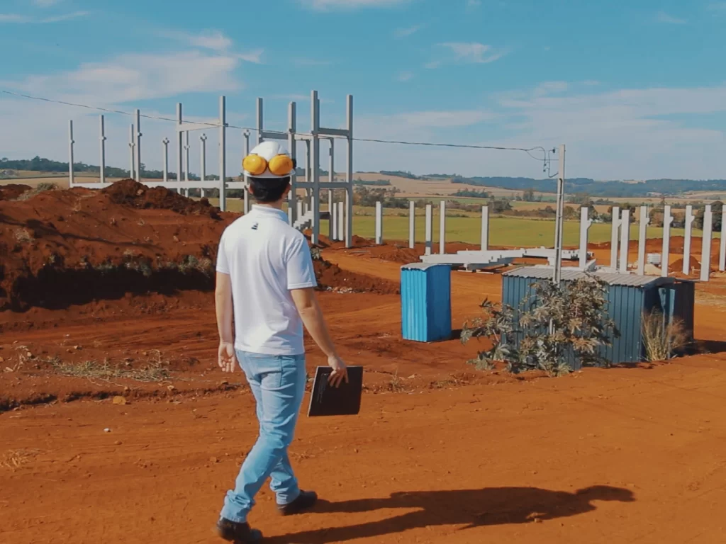 Engenheiro de capacete caminhando com prancheta pelo canteiro entre pilares pré-moldados