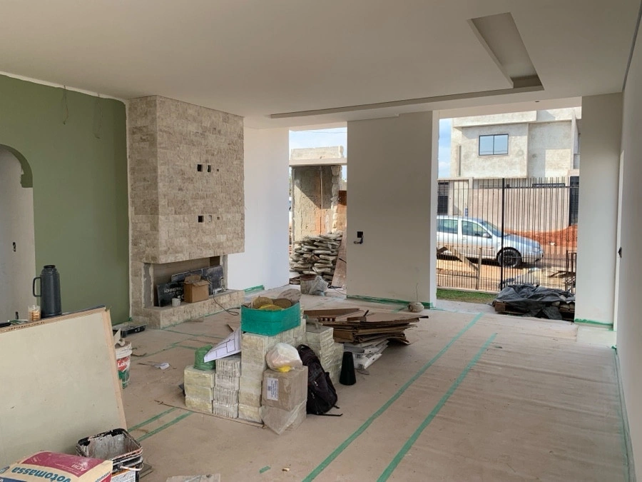 Sala de estar de residência em fase de acabamento, com lareira e coluna revestidas em pedra bege e forro de gesso rebaixado; piso coberto por proteção de obra e material de construção empilhado, e amplo vão de abertura para a área externa com portão