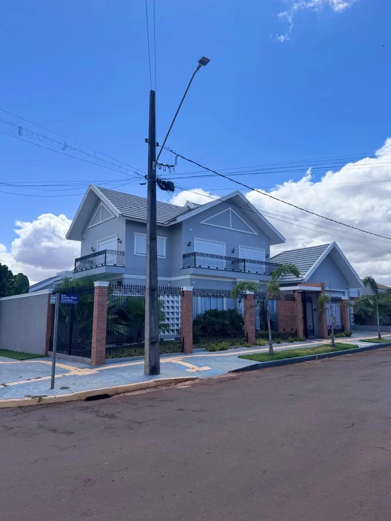 Sobrado de dois pavimentos entregue em esquina, fachada cinza-azulada com frontões e telhados em duas águas, sacada com gradil ornamental de ferro e divisa com pilares de tijolo aparente e gradil, com paisagismo concluído